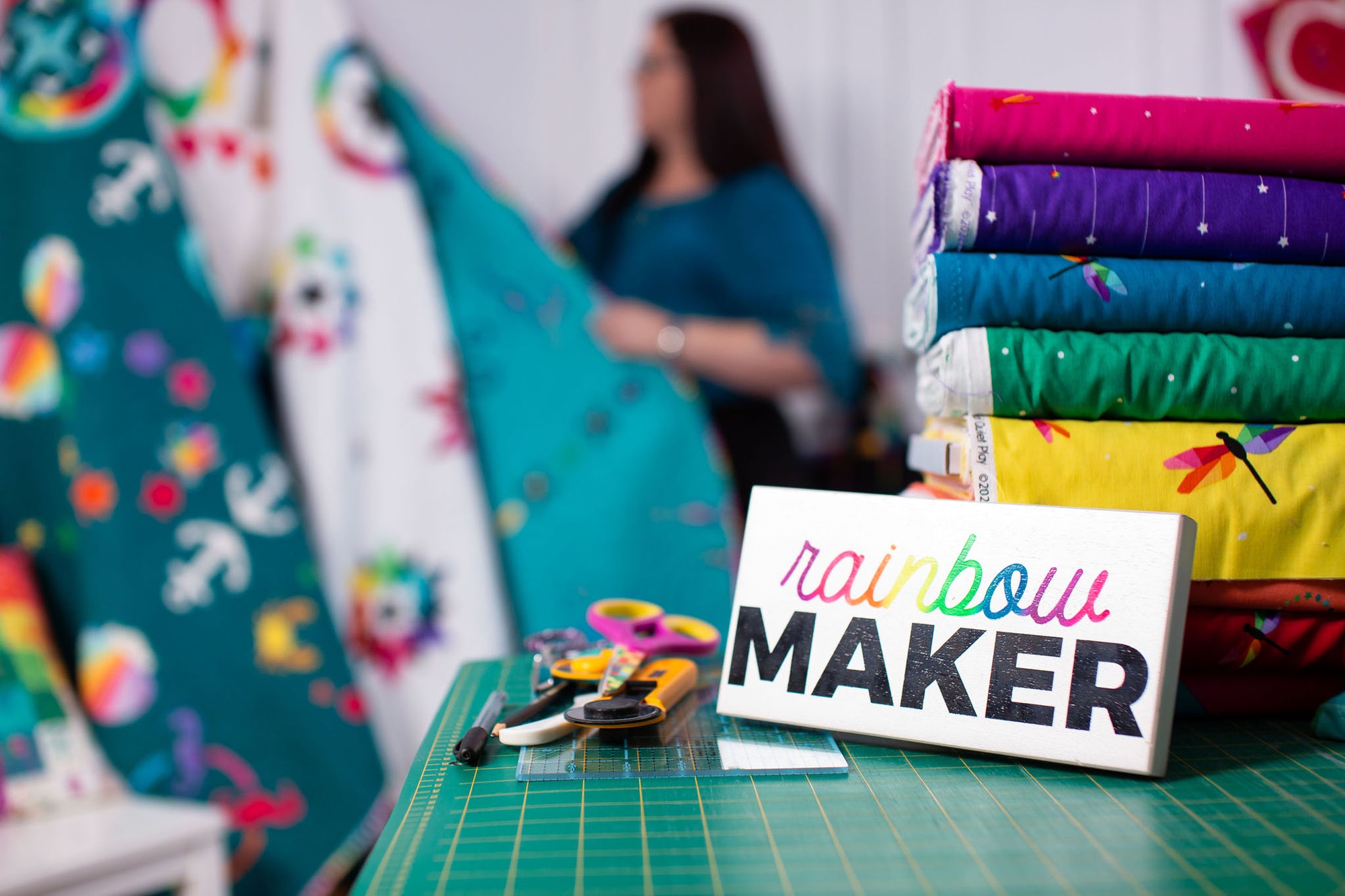 "Rainbow Maker" sign leaning against a stack of bright fabric rolls. In the background Kristy Lea is looking though FPP quilts.